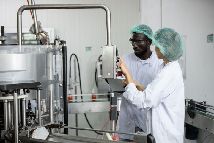 a man and a woman, wearing white lab coats and a head cover, operating a machinery